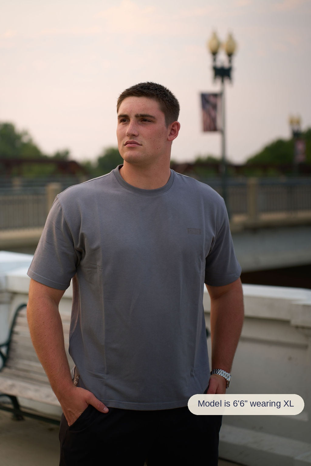 A man wearing a gray crewneck t-shirt and black pants standing outdoors on a bridge or walkway at sunset.