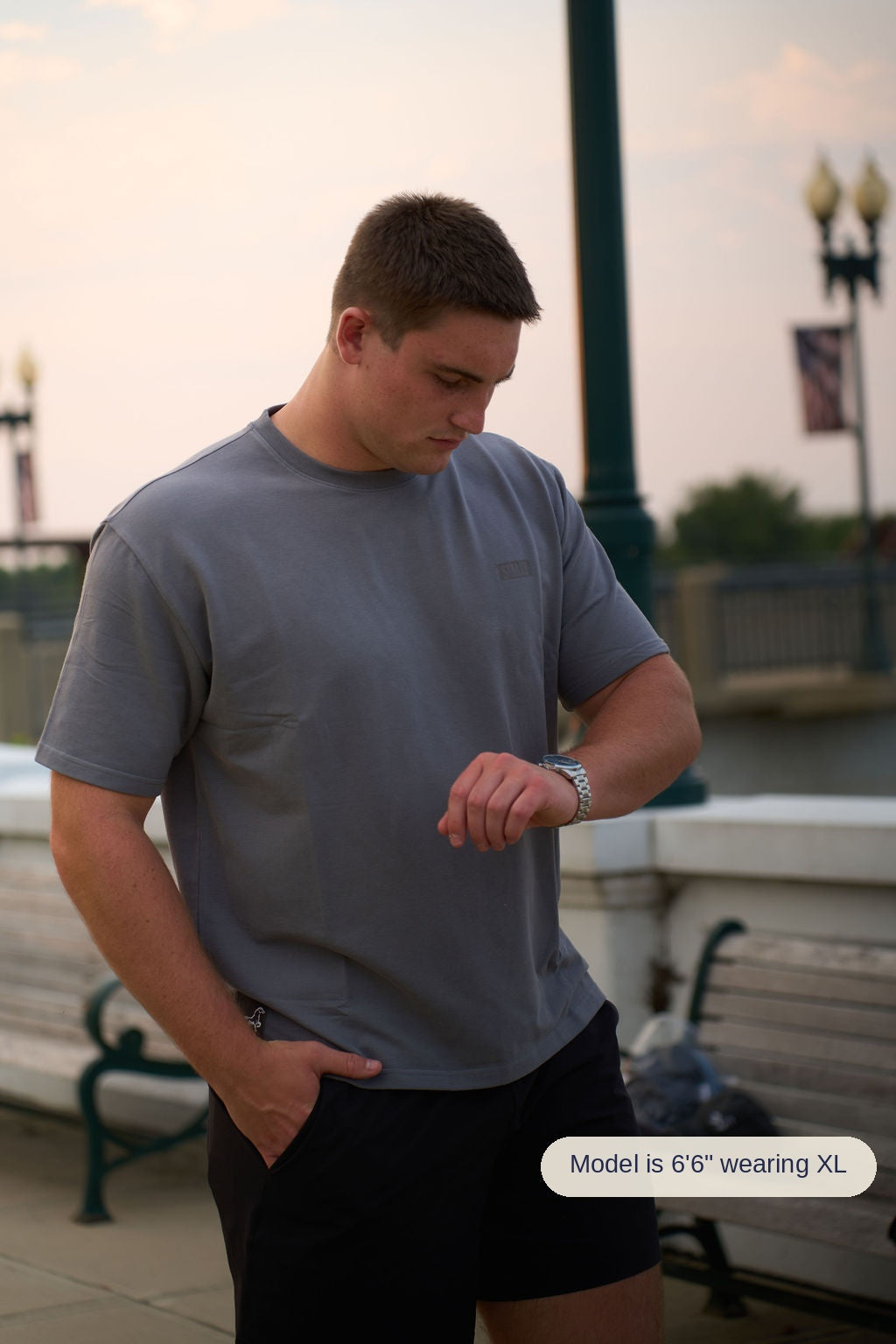 A man wearing a gray crewneck t-shirt and black shorts standing outdoors near a railing at sunset, looking down at his wristwatch.