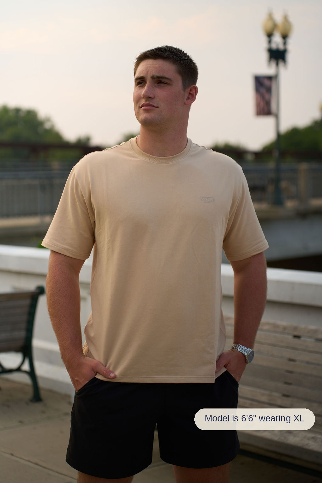A man wearing a saddlewood crewneck t-shirt and black athletic shorts, standing outdoors on a boardwalk with a bench and railing in the background.