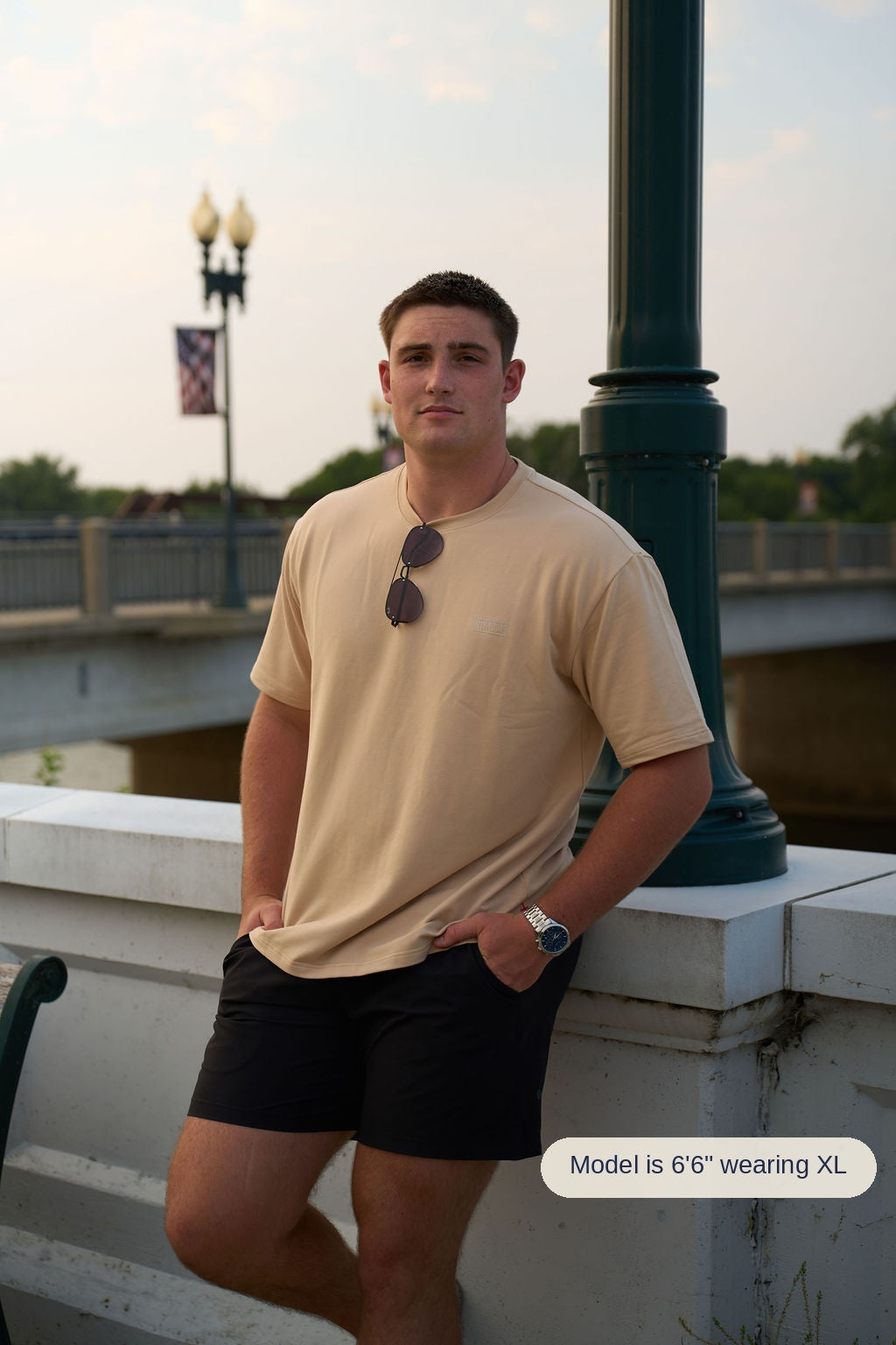 A man wearing a saddlewood crewneck t-shirt and black shorts stands leaning against a white stone wall next to a dark green post, with a bridge and street lamps in the background.