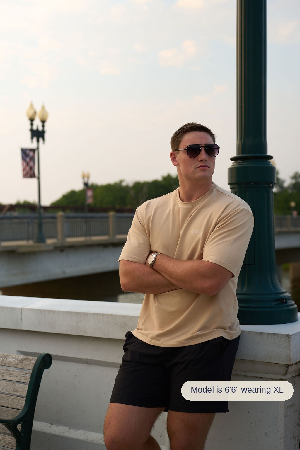 A man wearing a saddlewood crewneck t-shirt and black athletic shorts stands with arms crossed, leaning against a white stone wall next to a dark green lamp post outdoors.