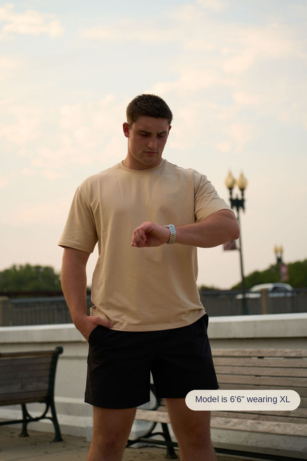 A man standing outdoors on a boardwalk looking down at his wrist while wearing a beige crewneck t-shirt and black athletic shorts.