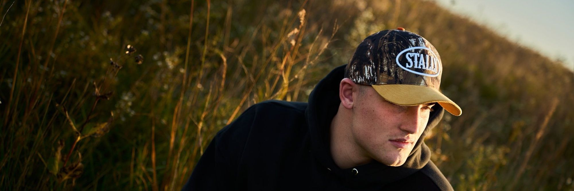 A man in a camo Stally oval-patch cap and black hoodie in tall grass at sunset