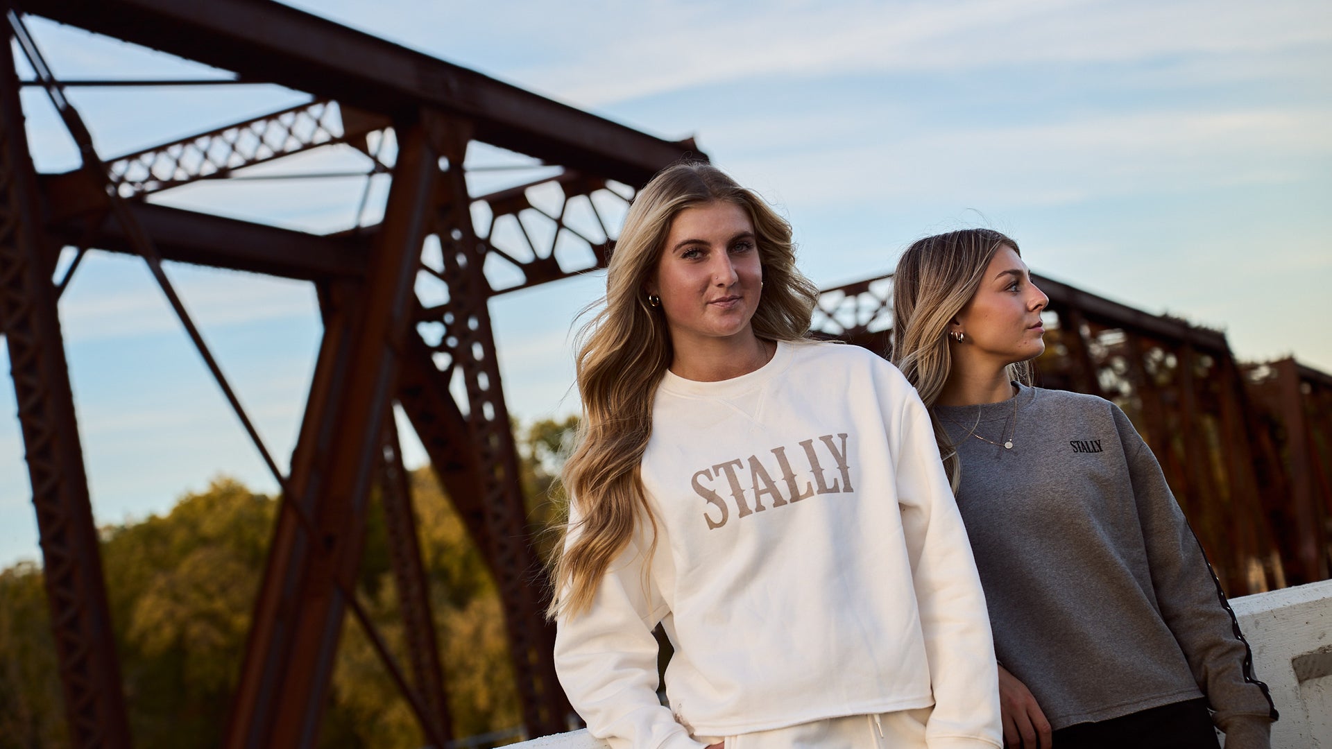Two women in Chalk White and Grey Heather Stally Range women's boxy crews on a rust-red iron bridge