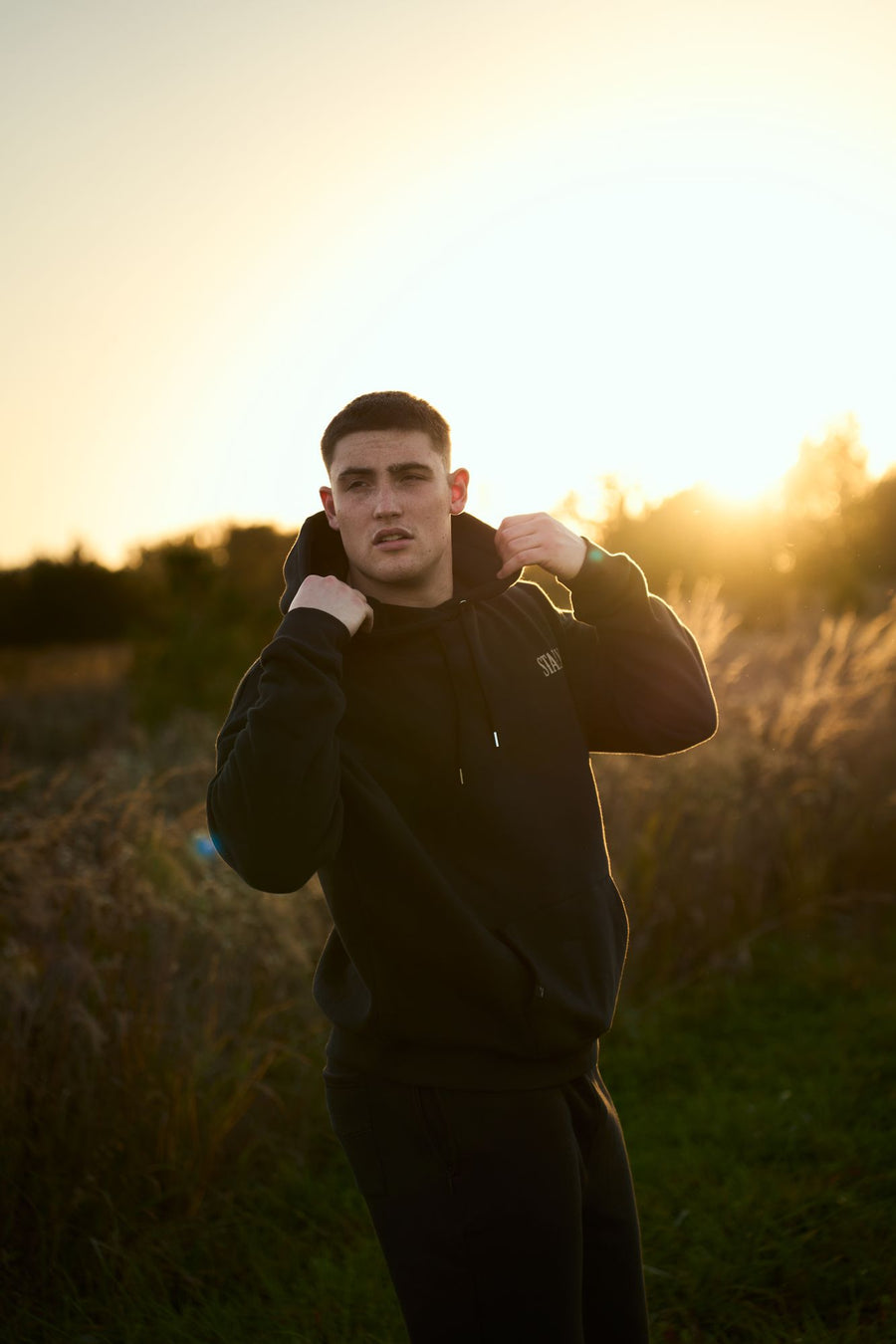 A man in a Coal Black Stally Draft hoodie pulling up the hood at sunset
